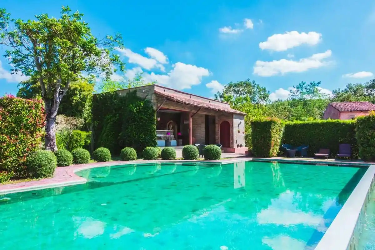 Linda piscina cercada por natureza exuberante, com uma casa ao fundo e céu azul claro. Ideal para relaxamento e lazer ao ar livre.