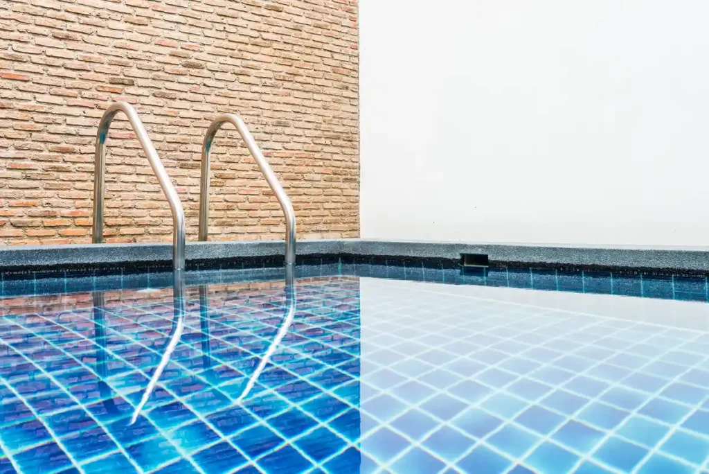 Piscina com degraus de entrada, paredes de azulejo azul e fundo com parede de tijolos e parede branca.