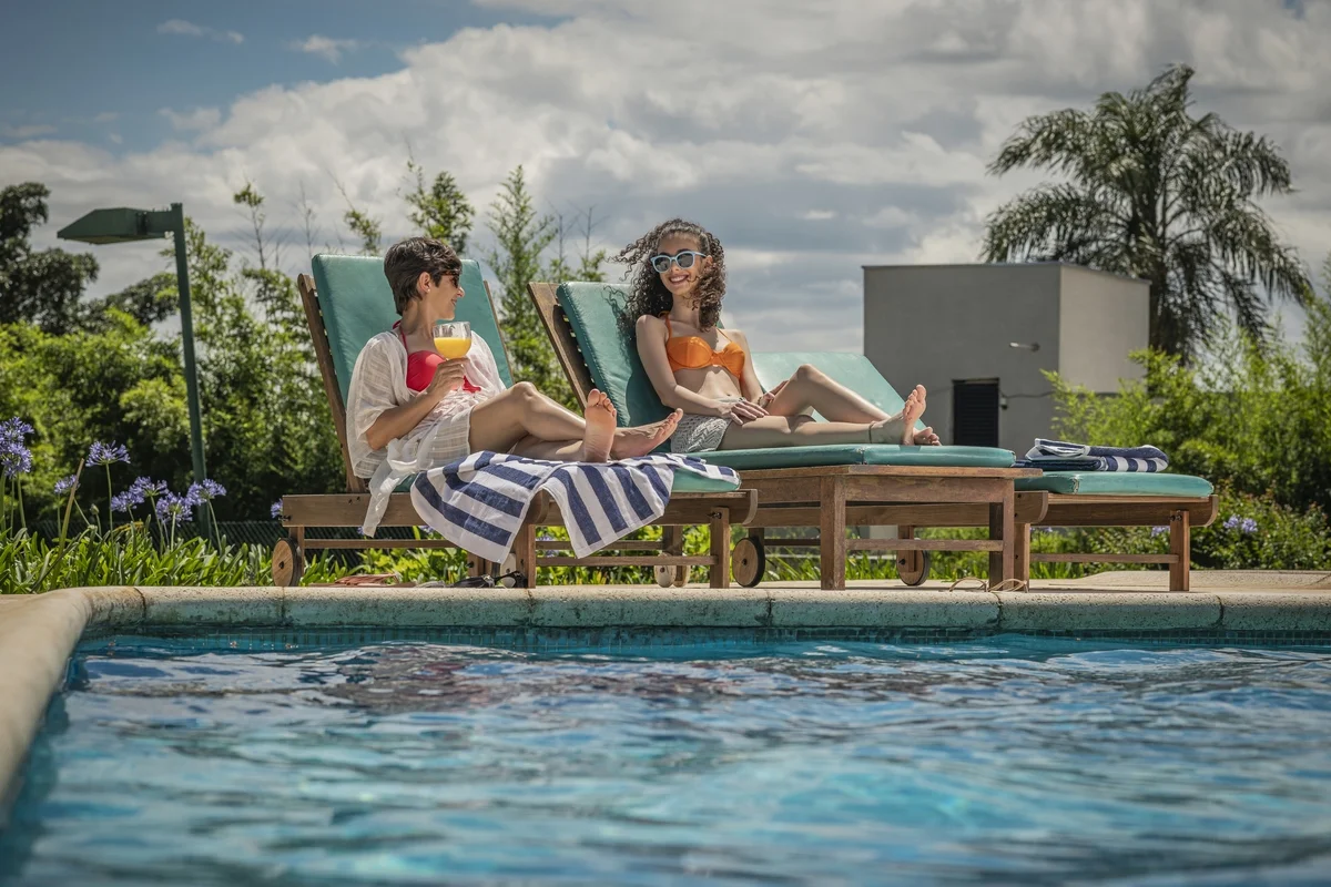 Duas mulheres relaxando em espreguiçadeiras ao lado de piscina com toalhas listradas e bebida