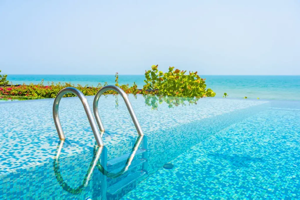 Vista de uma piscina de borda infinita com degraus prateados, refletindo o céu claro e o mar ao fundo, perfeita para um dia relaxante.