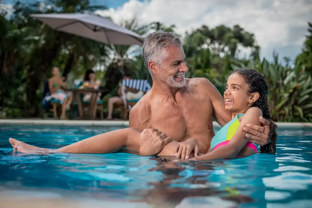 Homem e menina se divertindo na piscina ao ar livre em um dia ensolarado, com adultos e crianças ao fundo. Cenário tropical com árvores e céu claro.