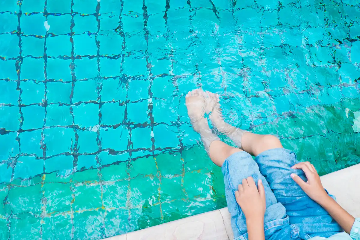 Pessoa segurando suas pernas enquanto relaxa à beira da piscina com água cristalina.