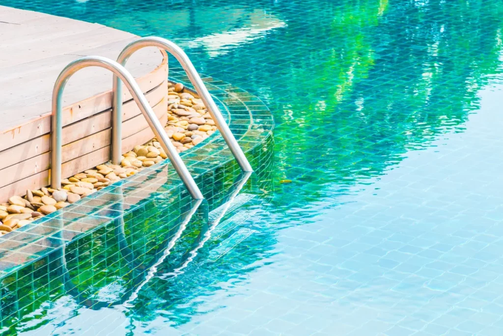 Escalera metálica en una piscina con borde de gresite verde y cubierta de madera con piedras decorativas
