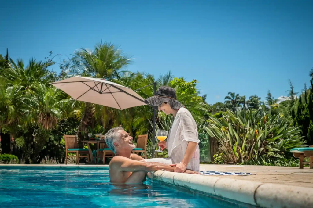 Una pareja feliz junto a la piscina bajo el sol, con un hombre mayor y una mujer joven disfrutando de unas copas en un entorno tropical con muchas plantas y adornos.