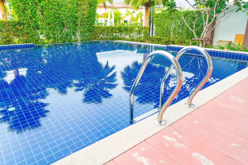 Piscina residencial com azulejos azuis e escada de metal, cercada por plantas verdes e palmeiras refletidas na água