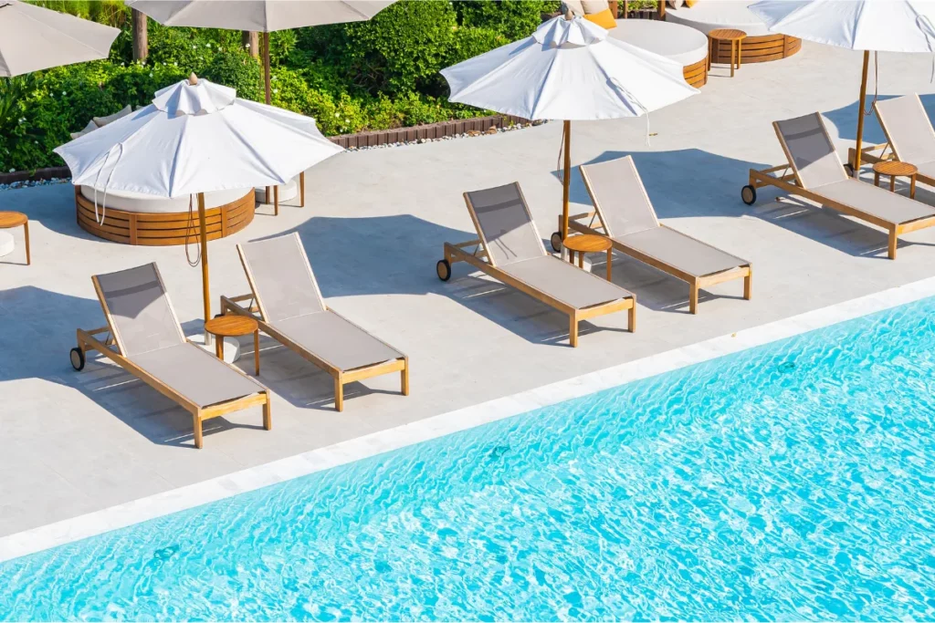 Tumbonas de madera con tela beige y sombrillas blancas junto a una piscina azul en una zona exterior con suelo claro y vegetación de fondo.