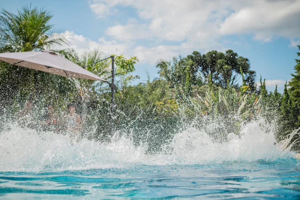 Água espirrando numa piscina com plantas tropicais e um grande guarda-sol ao fundo, sob um céu parcialmente nublado.