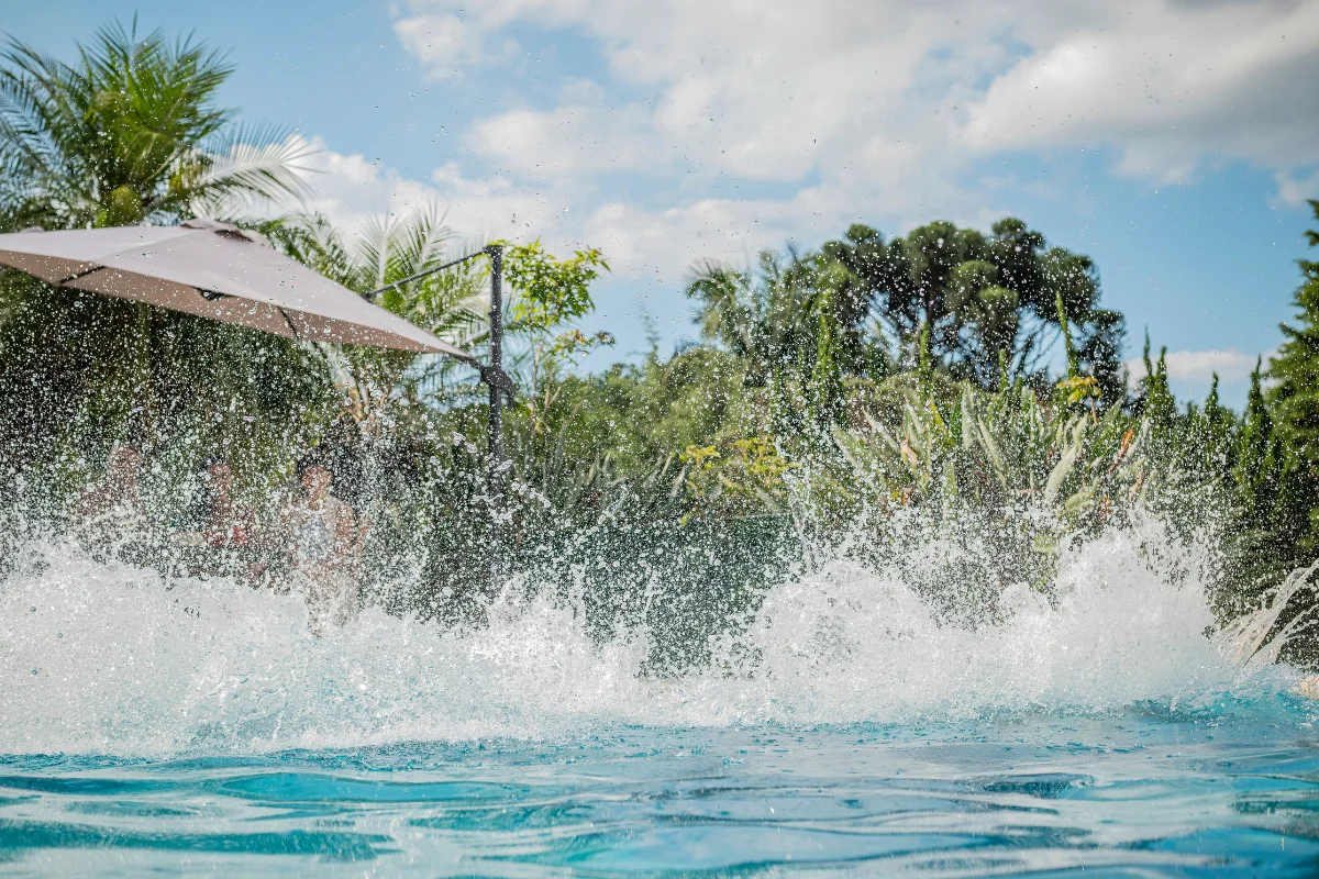 Água espirrando numa piscina com plantas tropicais e um grande guarda-sol ao fundo, sob um céu parcialmente nublado.